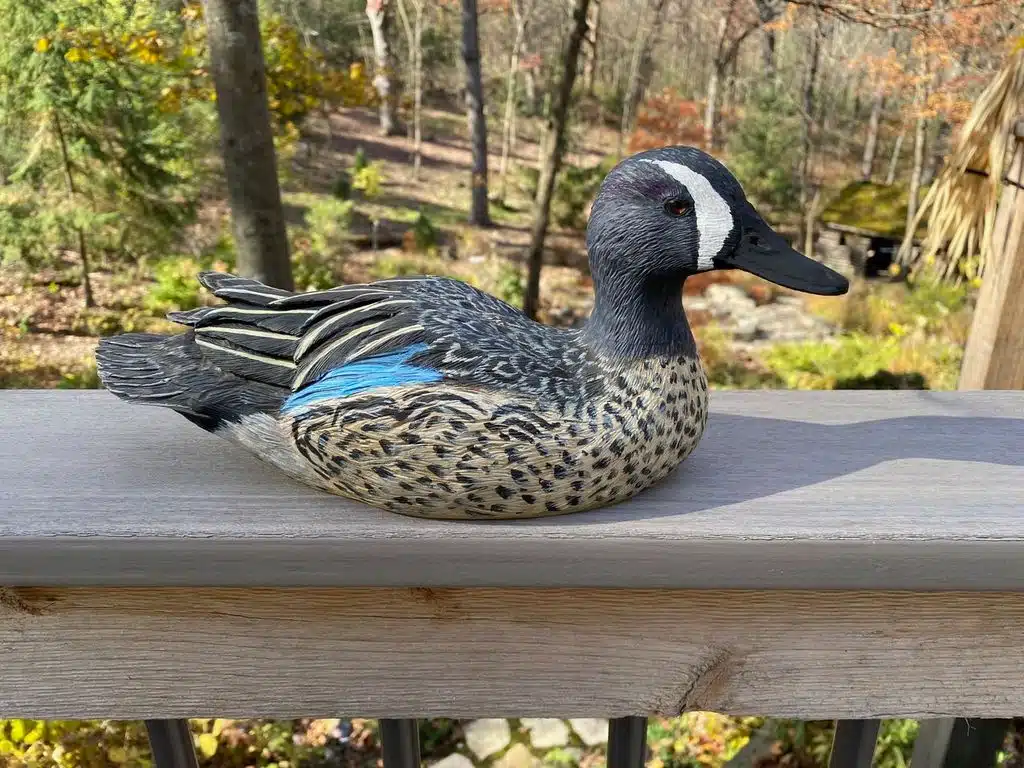 A detailed wooden duck decoy on a railing in a wooded area.